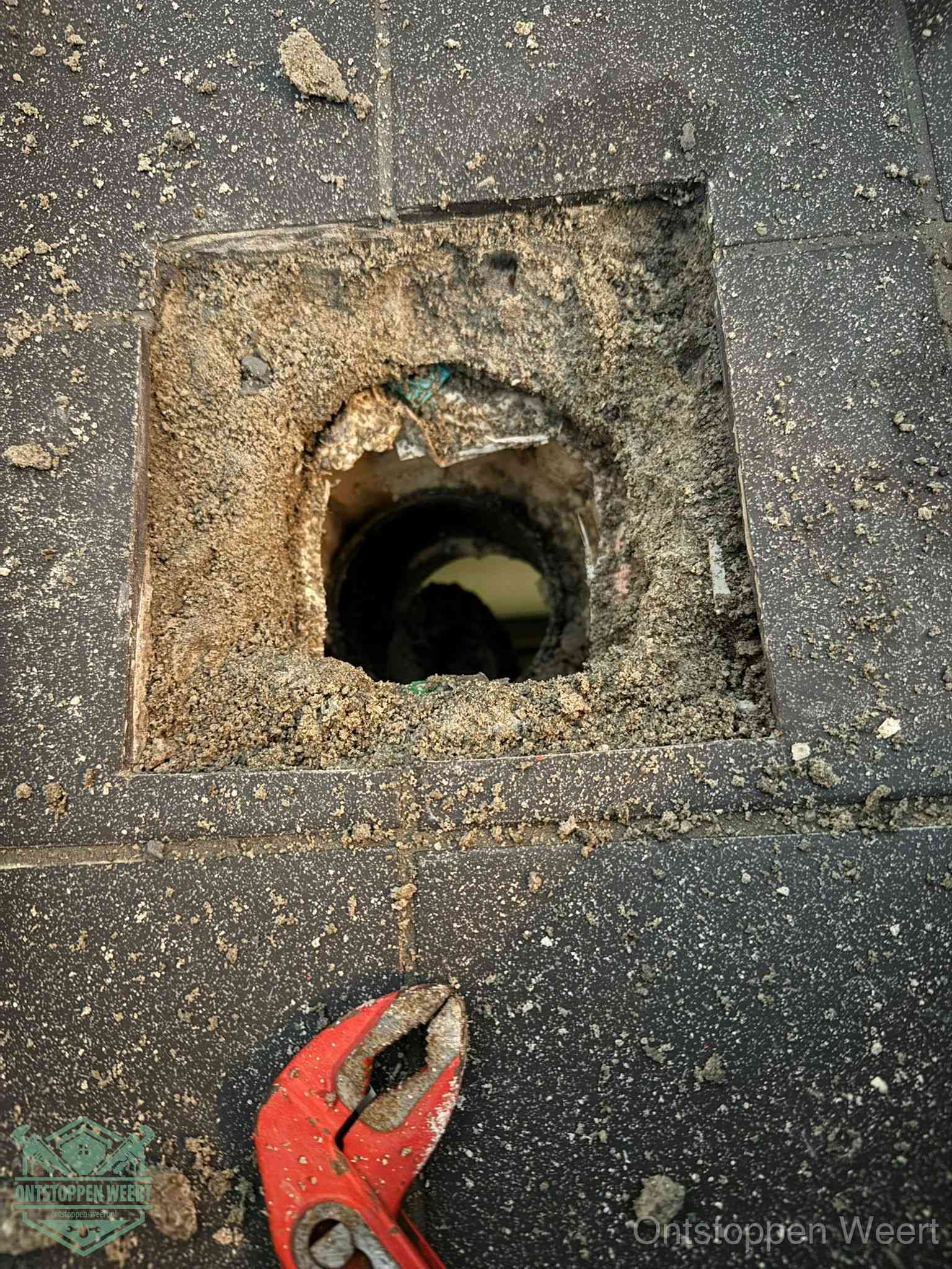 Een geopend rioolputje in een betontegelvloer met losgemaakt rooster en een zichtbare ronde afvoerbuis, het achterhalen waar de afvoerverstopping zit in Weert