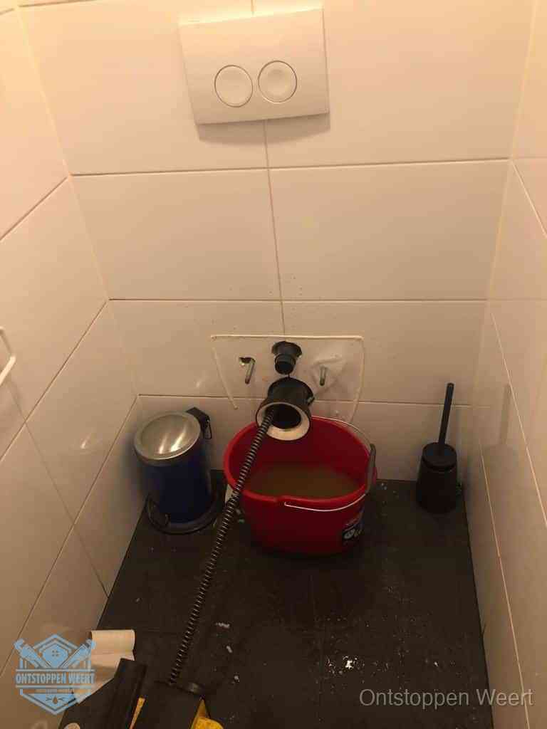 Een ontstopte wand-wc met verwijderde toiletpot waar een ontstoppingsveer in de wanduitgang verdwijnt, het achterhalen waar de verstopping zit in Weert