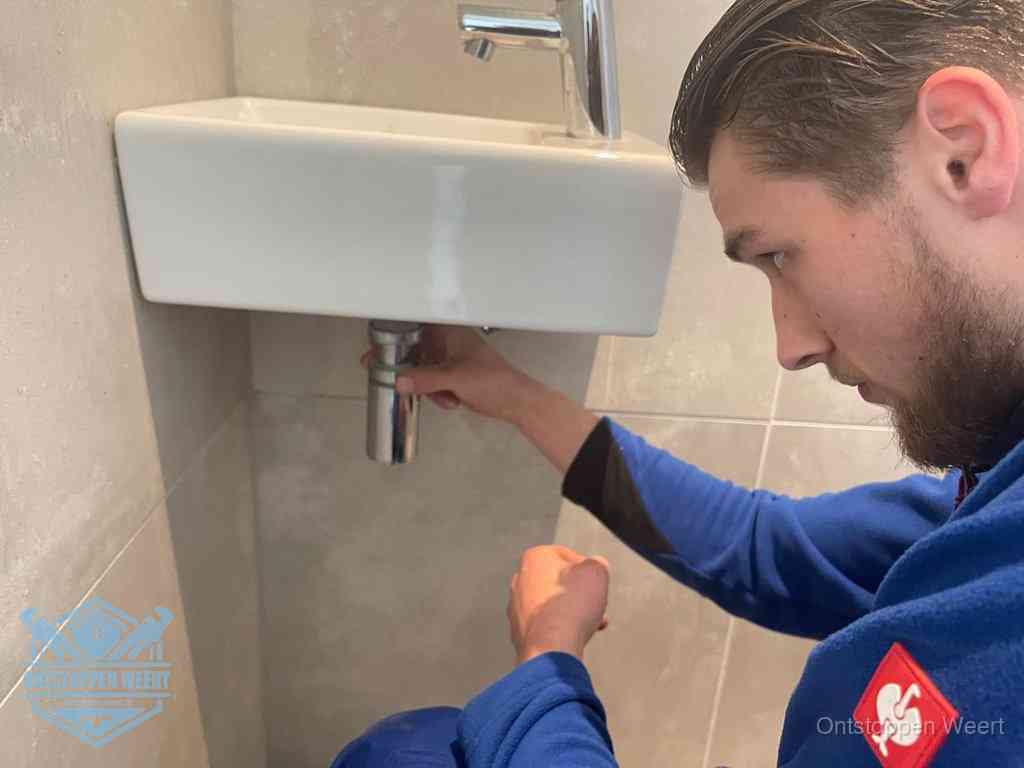 Een monteur draait met de hand de zwanenhalssifon onder een fonteinwasbak in de toiletruimte los om de bron van rioollucht in Weert te vinden