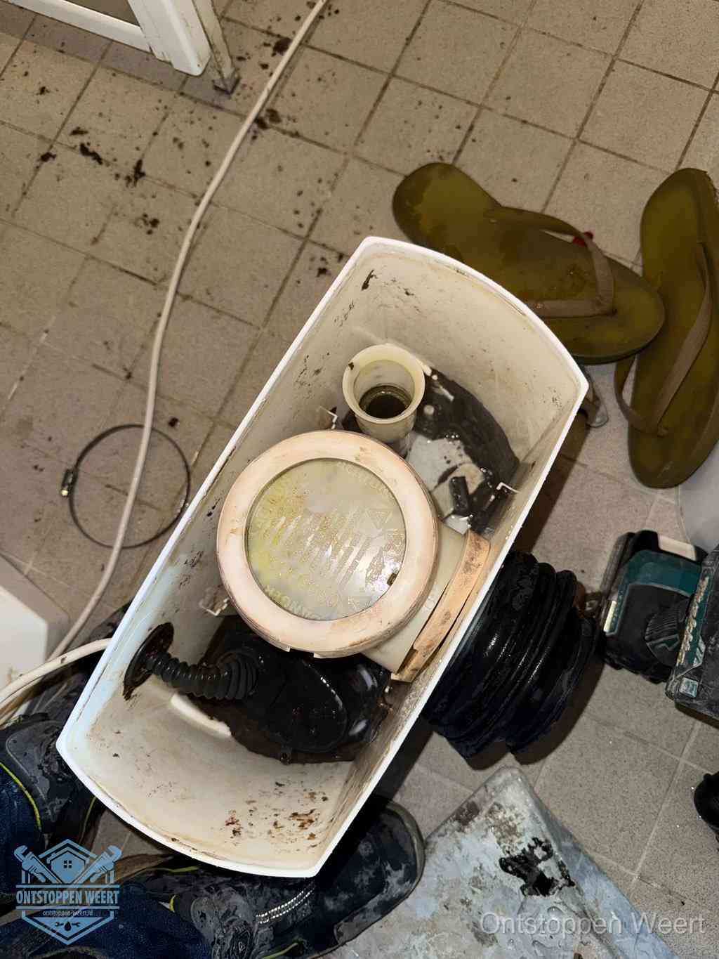 Een gedemonteerde toiletpot op de vloer met ontstoppingsveer en gereedschap ernaast, het verwijderen van een vastzittend voorwerp uit de wc in Weert