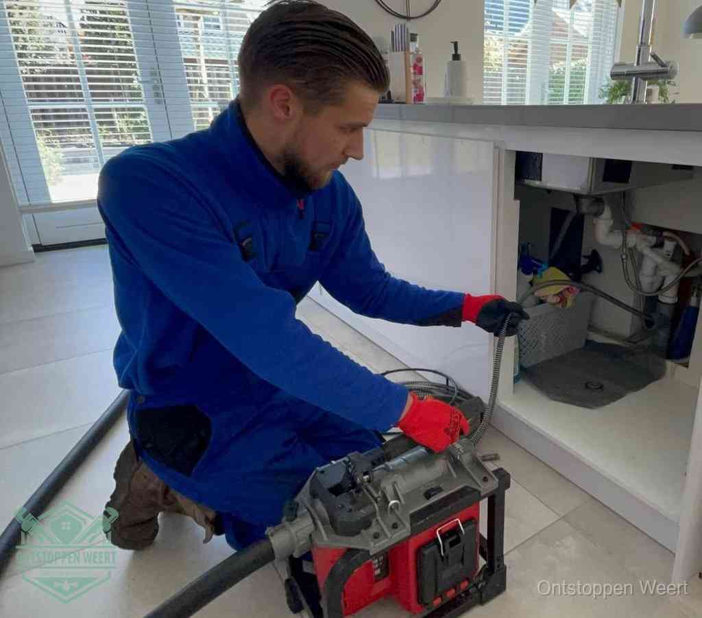 Een monteur met rode handschoenen knielt in een keuken en voert een elektrische ontstoppingsveer in tijdens het professioneel afvoer ontstoppen in Weert